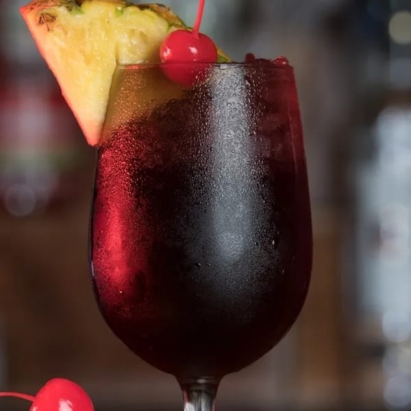 A vibrant red Jamaican drink in a wine glass, garnished with a pineapple slice and cherry.