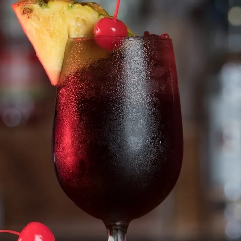 A vibrant red Jamaican drink in a wine glass, garnished with a pineapple slice and cherry.