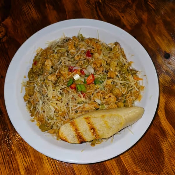A plate of Jamaican-style pasta with shredded cheese, chopped vegetables, and grilled bread on a wooden table.