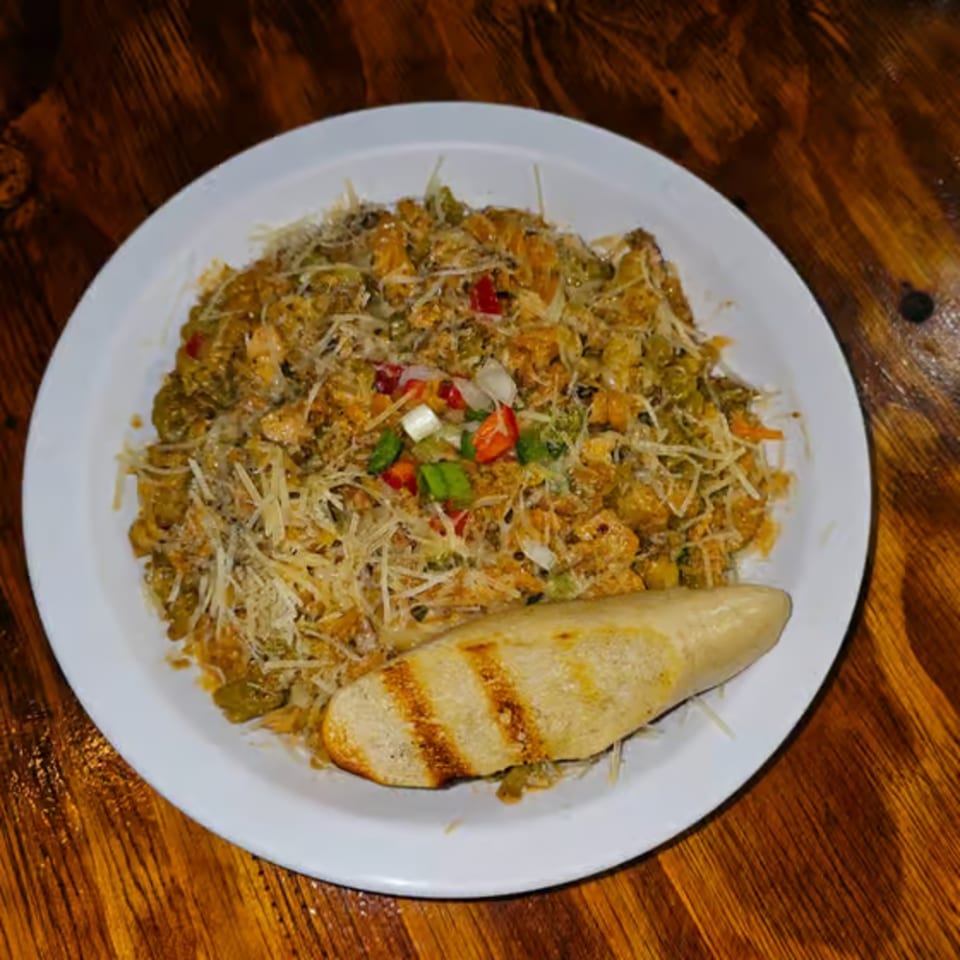 A plate of Jamaican-style pasta with shredded cheese, chopped vegetables, and grilled bread on a wooden table.